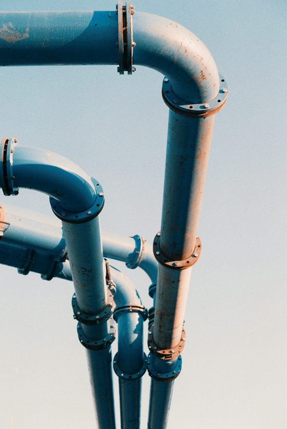 Upward-looking photo of several large light-blue industrial pipelines with curved joints and bolted flanges against a clear pale-blue sky.